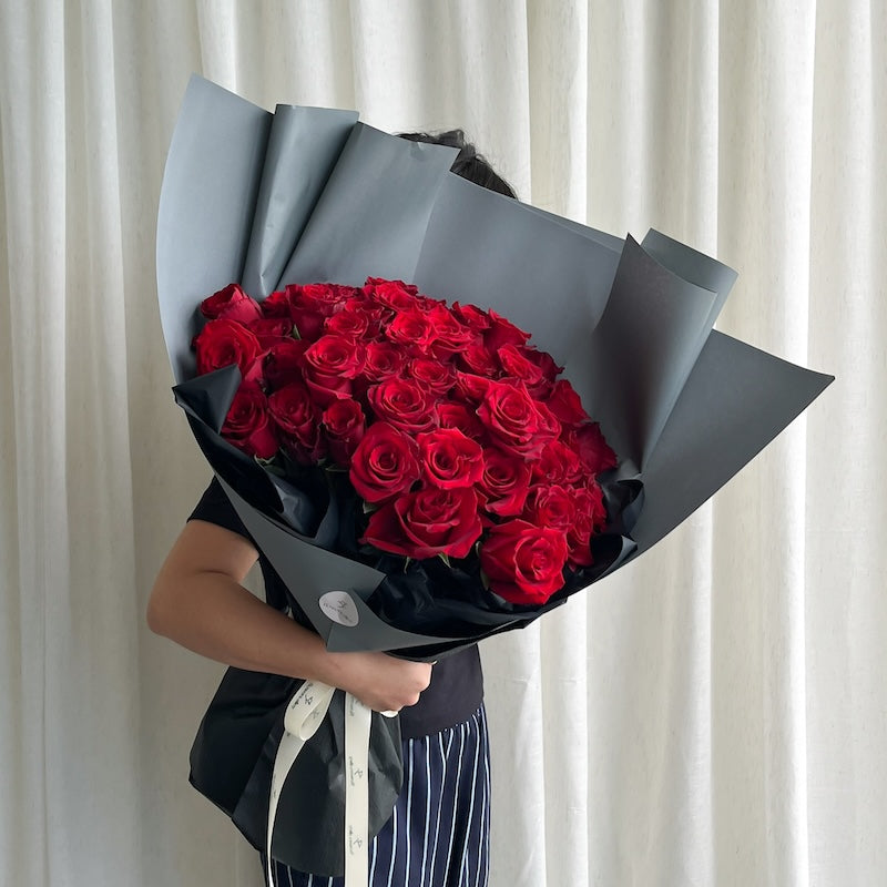 Bouquet of red roses wrapped in black paper held by a person against a white curtain background